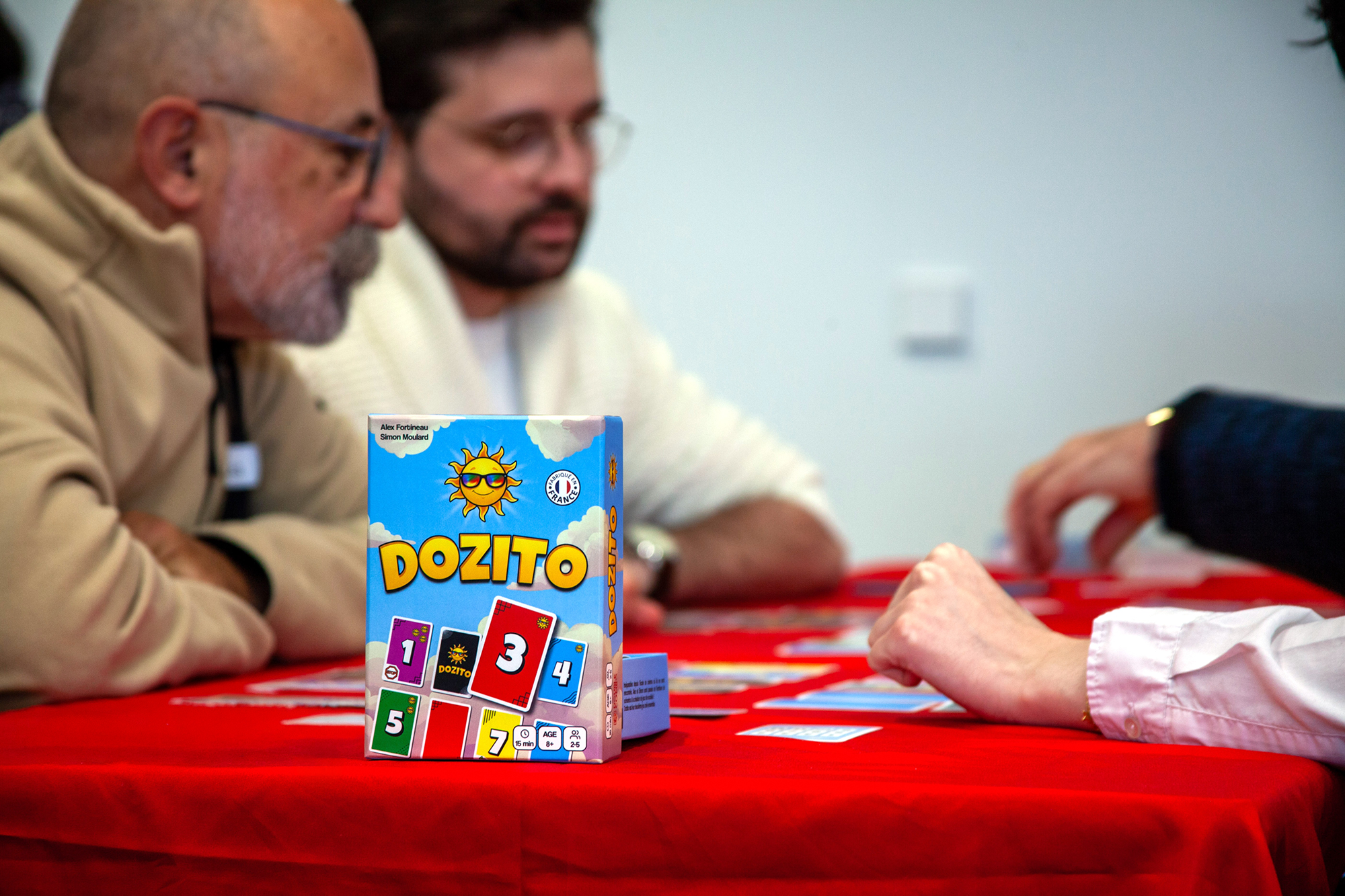 Une boite de dozito sur une table rouge, devant deux personnes qui jouent à Dozito.
